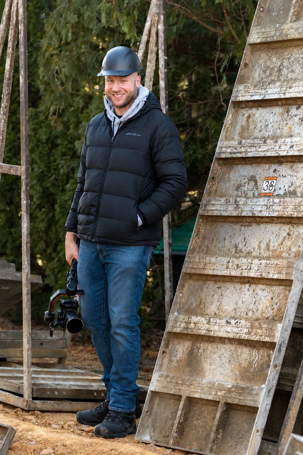 Ryan Samuels on a job site in a hard hat, holding a camera on a gimbal beside concrete forms