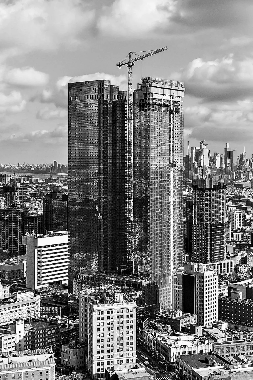 Black and white view of two glass towers rising in Jersey City, a tower crane between them and Manhattan beyond