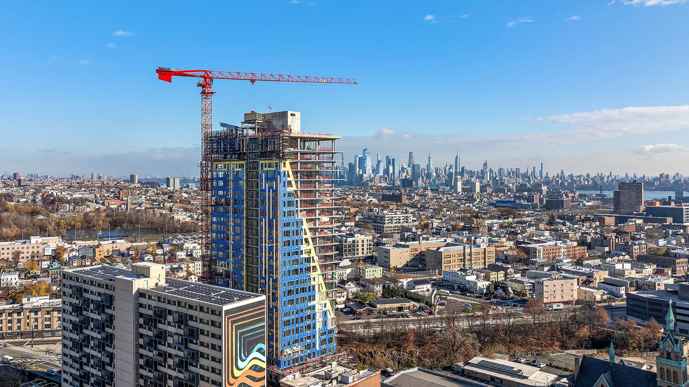 Red crane atop a rising tower with a sloped glass facade, Manhattan on the horizon