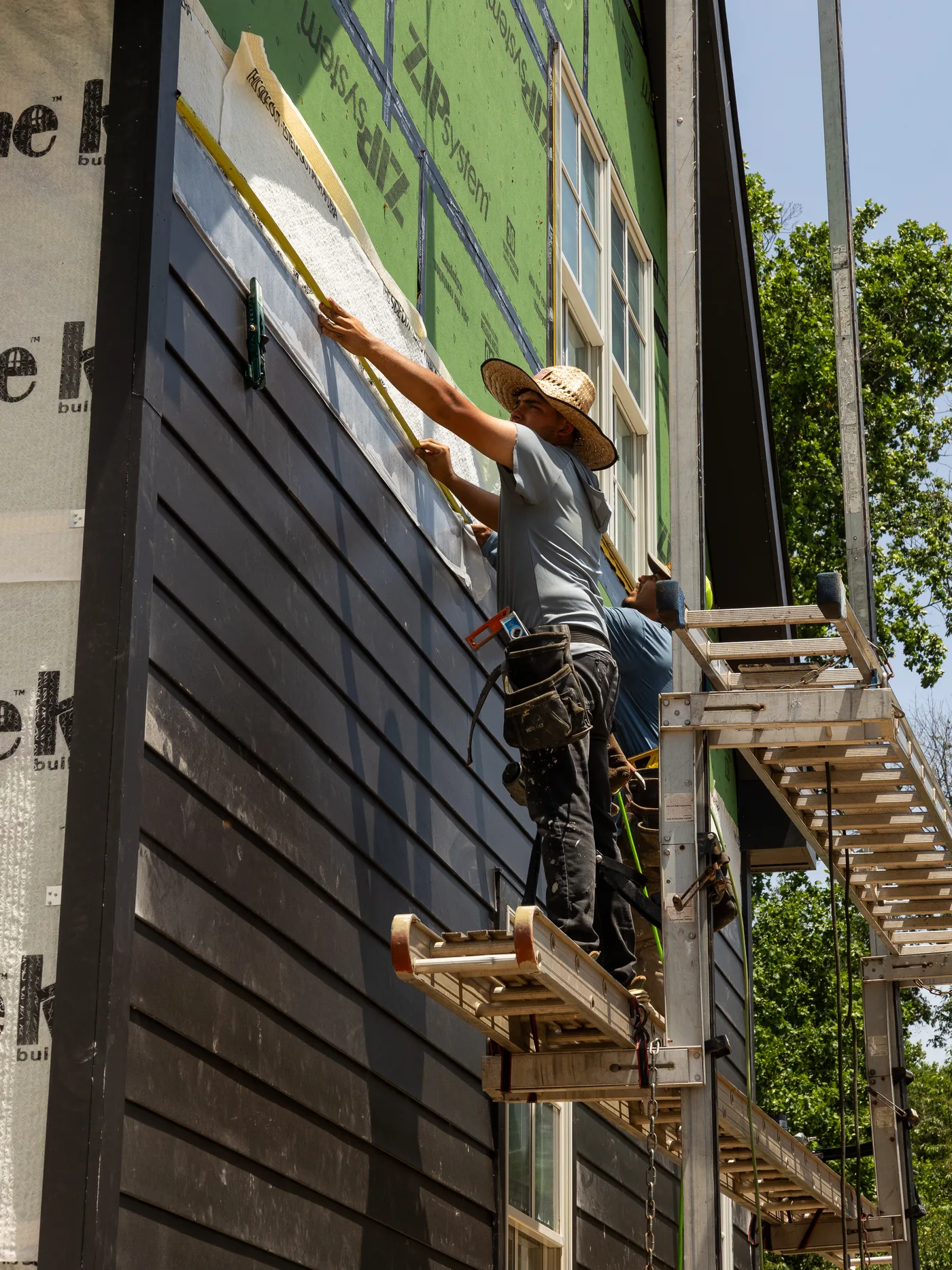 Installer on a scaffold nailing dark siding over house wrap