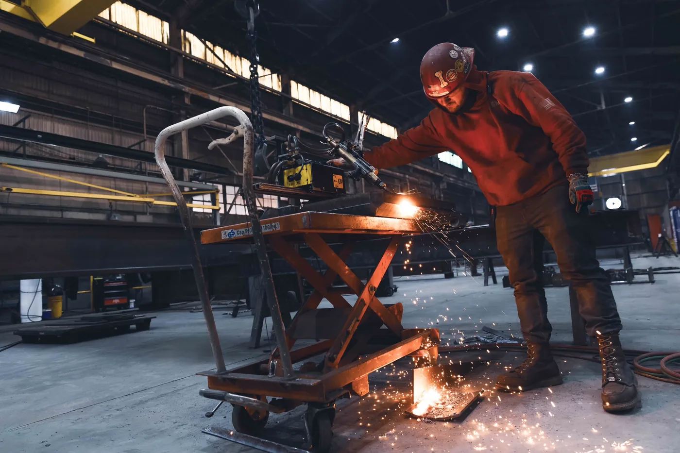 Worker in a red jacket grinding steel on a lift table as sparks fall to the floor