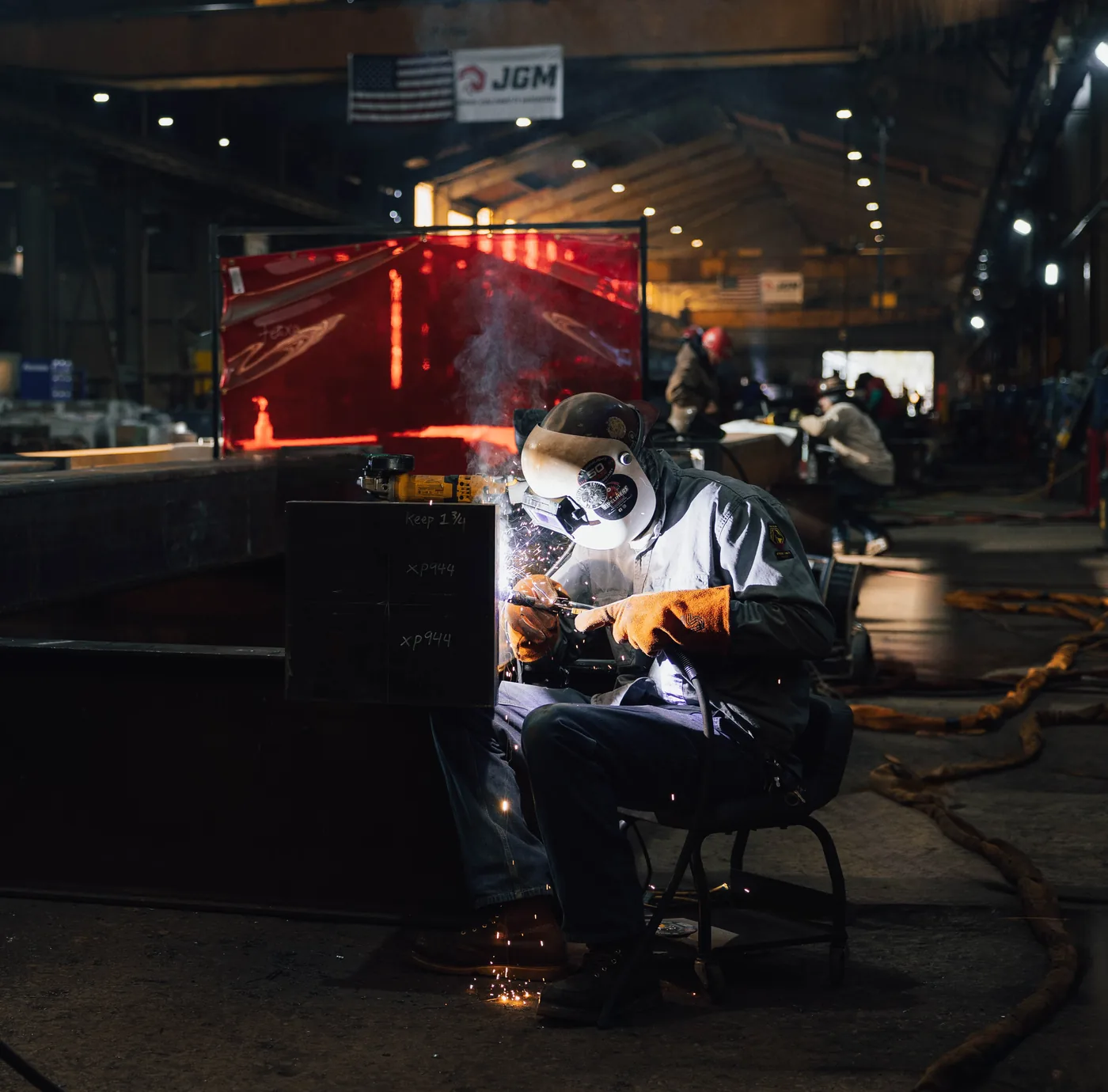 Seated welder at work beneath a large American flag in the fabrication shop