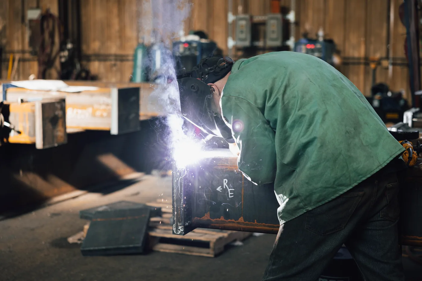 Welder in a green jacket bent over a steel assembly, arc light flaring across the beam