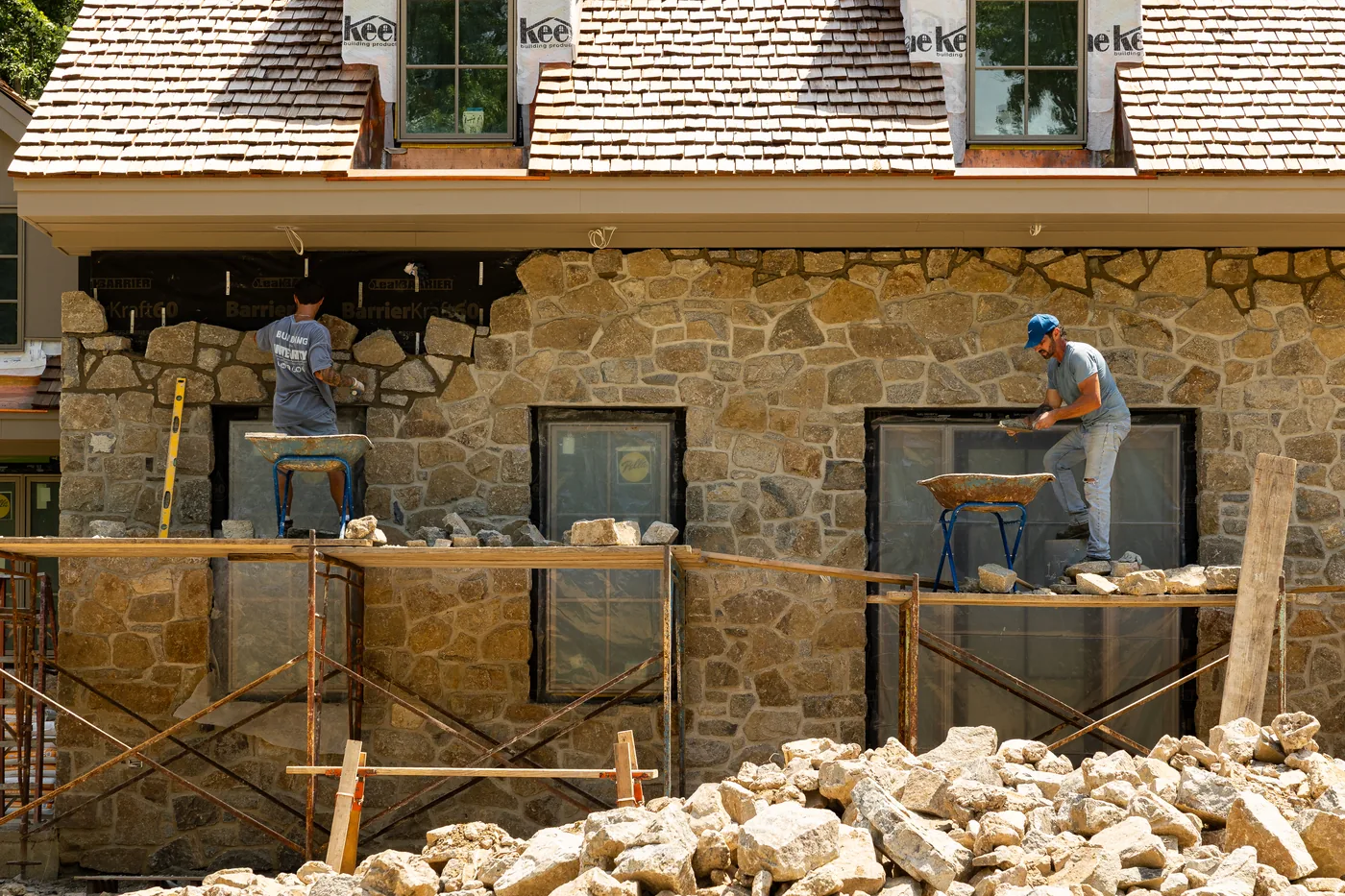 Two masons on a scaffold setting fieldstone around the windows of a new stone facade