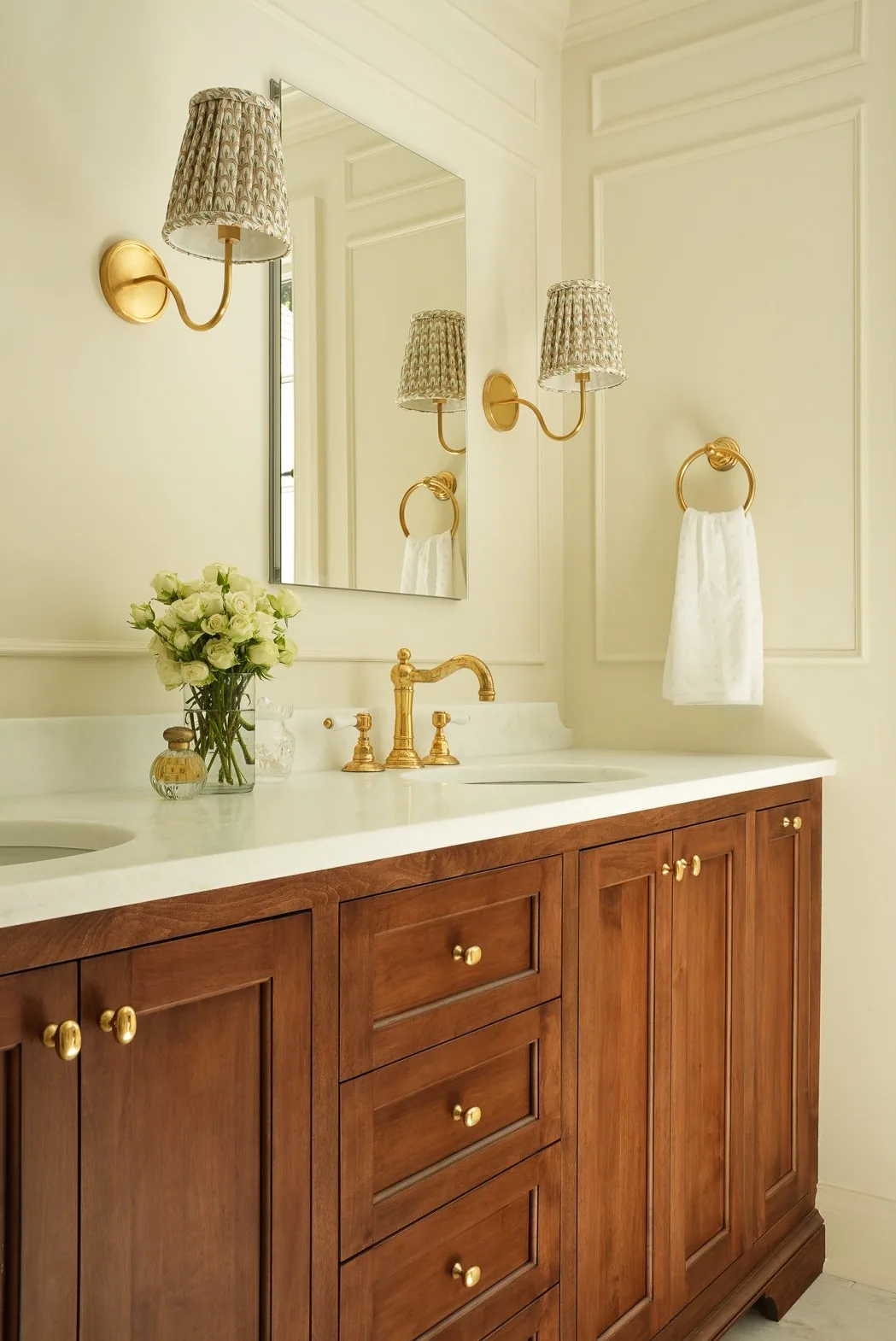 Walnut double vanity with brass fixtures beneath pleated shade sconces and paneled walls