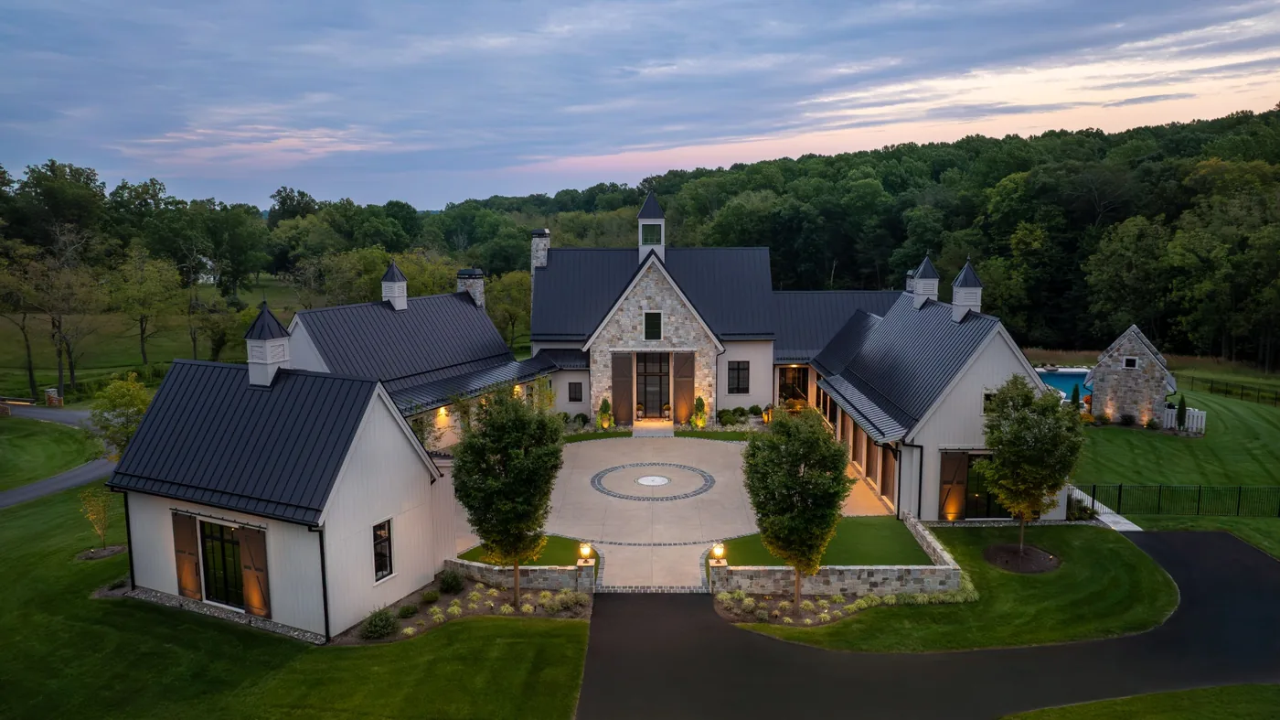 Stone and board and batten farmhouse estate at dusk, its courtyard and cupolas lit warmly