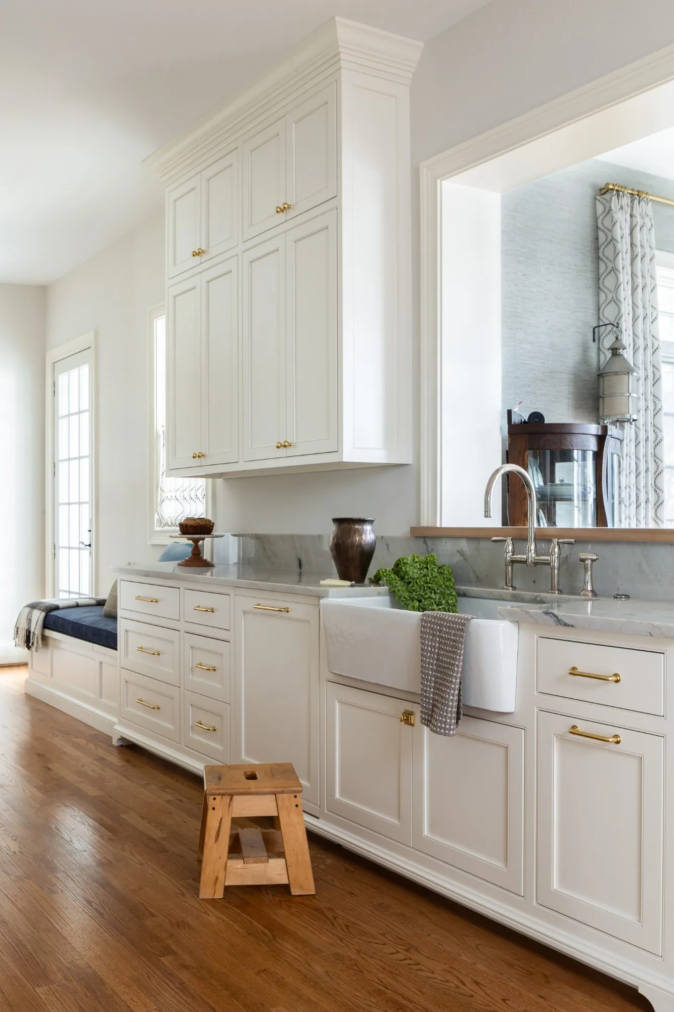 White inset cabinetry with brass pulls, a farmhouse sink, and a window seat beside the pass through