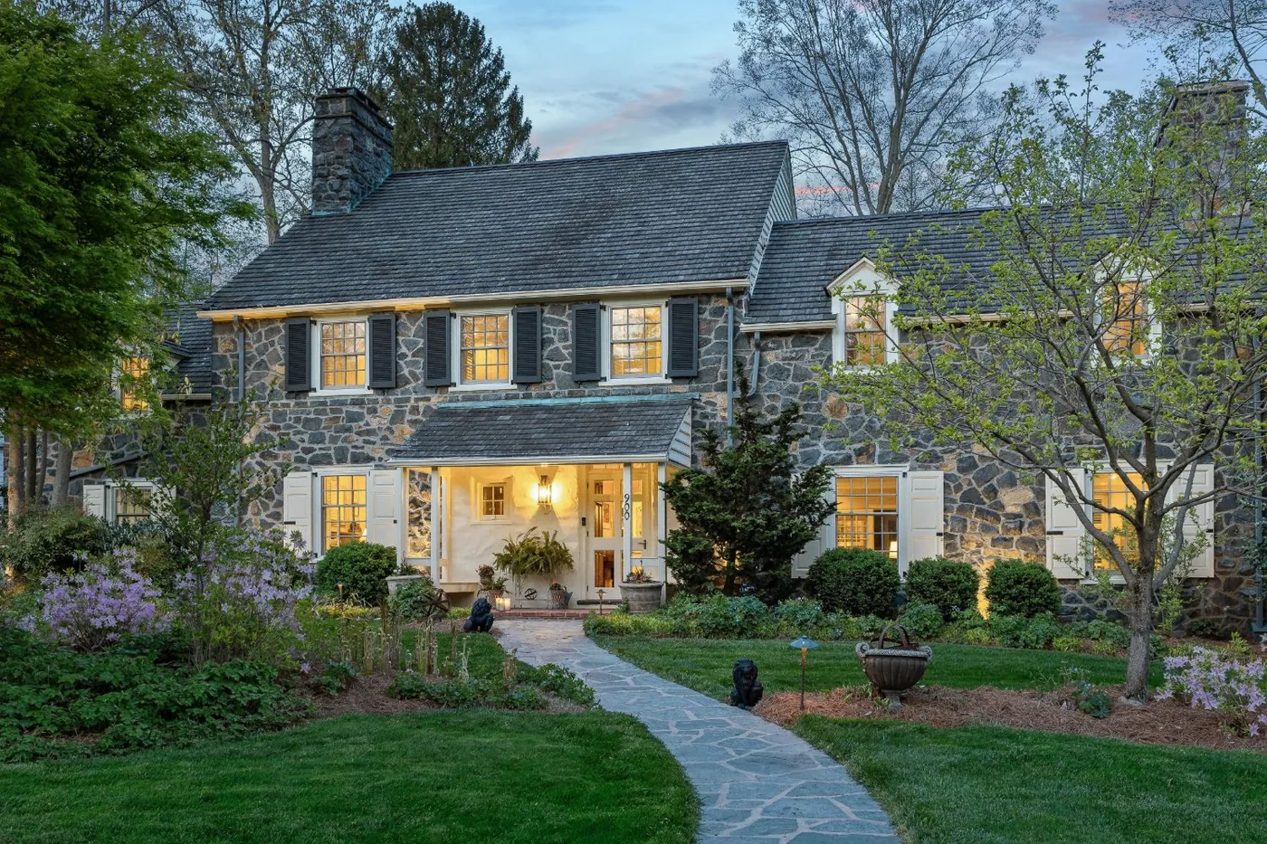 Fieldstone colonial at blue hour, warm windows and a lit porch at the end of a curving flagstone path